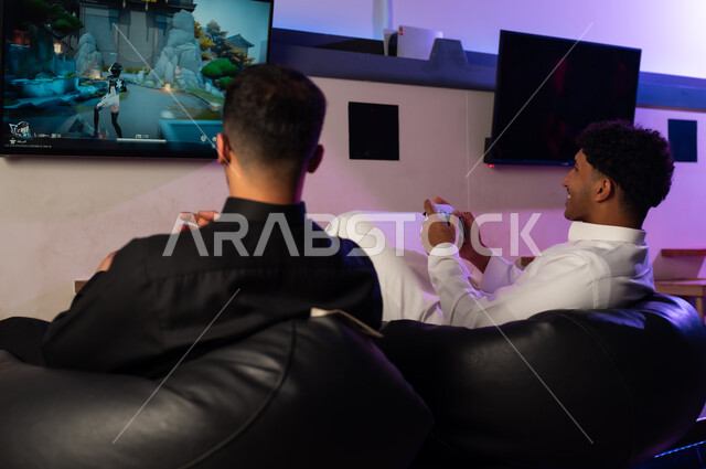 Holding the remote control of the video game, two Arab Gulf men from Saudi Arabia, one wearing a white thobe and the other a black thobe, are sitting happily while playing, background image, enjoyable youthful atmosphere.