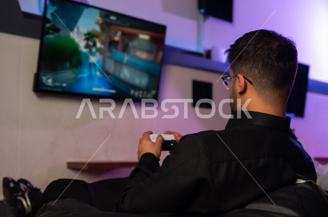 رجل عربي خليجي سعودي يرتدي الثوب الاسود يجلس على المقعد , النظر للشاشة بتركيز و اللعب بشغف , تصوير خلفي , انشطة ترفيهية شبابية في مركز الالعاب الالكترونية
