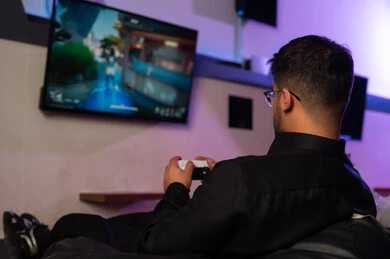 A Saudi Arabian Gulf man wearing a black thobe is sitting on a chair, looking at the screen with focus and playing passionately, captured from behind, engaging in youth recreational activities at an electronic games center.