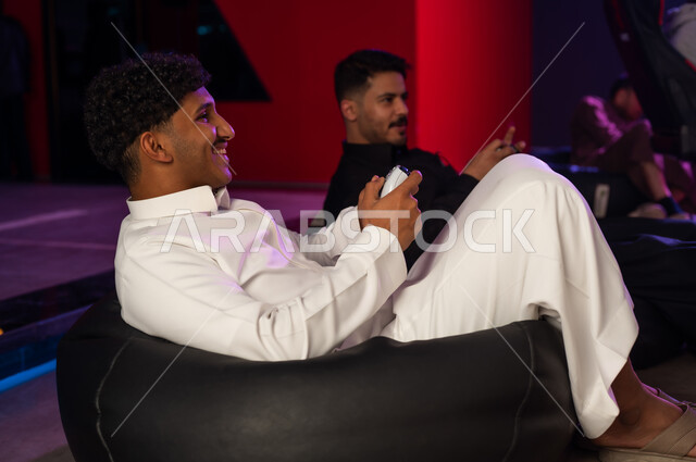 لحظات ممتعة بين الاصدقاء , رجل عربي خليجي سعودي يرتدي الثوب الابيض يجلس على الكرسي المريح , شاب عربي خليجي سعودي يرتدي الثوب الاسود ينظر الى الشاشة , انشطة شبابية مرحة في مركز الالعاب الالكترونية