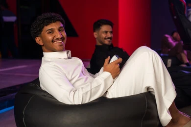 A Saudi Arabian Gulf man wearing a white thobe is looking at the camera with a smile, a Saudi Arabian Gulf man wearing a black thobe is happily holding a game controller, moments of fun and enjoyment with friends.