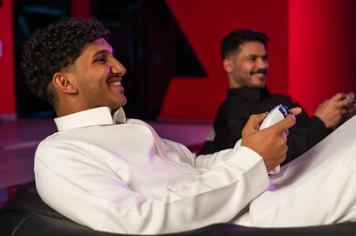 النظر الى الشاشة بابتسامة وبداية اللعبة الممتعة , رجل عربي خليجي سعودي يرتدي ثوب ابيض يجلس على المقعد الاسود المريح , رجل عربي خليجي سعودي يرتدي الثوب الاسود يمسك جهاز التحكم باللعبة الالكترونية , ايماءات السعادة و البهجة