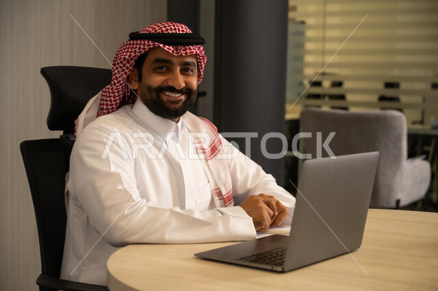 إدارة شؤون الأعمال وتنظيمها، استخدام جهاز تقني حديث ومتطور، مهنة ووظيفة ادارية مكتبية، موظف عربي خليجي سعودي يرتدي الثوب التقليدي يقوم بالعمل على الحاسوب المحمول داخل المكتب، الشركات والمكاتب في المملكة العربية السعودية