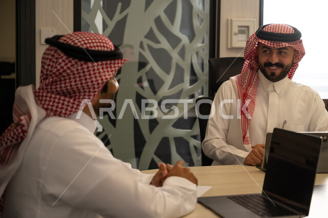 الاجتماعات الإدارية والتواصل المهني داخل الشركات، موظف عربي خليجي سعودي يرتدي الثوب و الشماغ يجلس على طاولة اجتماع يتحدث مع زميله أثناء جلسة عمل رسمية، استخدام الحاسب المحمول لمتابعة الأعمال ومناقشة المهام، مفهوم التعاون الوظيفي واتخاذ القرارات