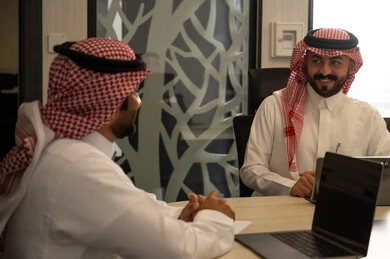 الاجتماعات الإدارية والتواصل المهني داخل الشركات، موظف عربي خليجي سعودي يرتدي الثوب و الشماغ يجلس على طاولة اجتماع يتحدث مع زميله أثناء جلسة عمل رسمية، استخدام الحاسب المحمول لمتابعة الأعمال ومناقشة المهام، مفهوم التعاون الوظيفي واتخاذ القرارات