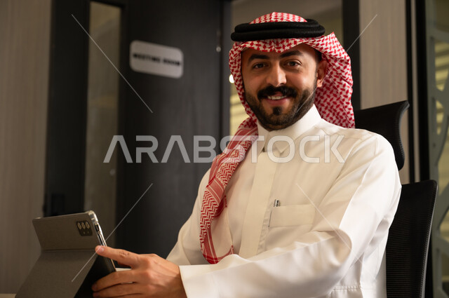 Accomplishing tasks using a smart device, an Arab Gulf Saudi man wearing a white thobe and a ghutrah sits inside the office holding a tablet, looking at the camera with a joyful expression.
