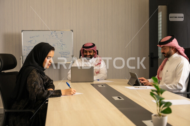 مفهوم التخطيط والتنظيم الإداري, اجتماع فريق عمل لمناقشة المهام, موظفون عربيون خليجيون سعوديون يرتدون الزي التقليدي يستخدمون الحاسب المحمول والأجهزة اللوحية داخل غرفة الاجتماع، متابعة سير العمل