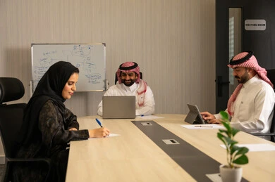 مفهوم التخطيط والتنظيم الإداري, اجتماع فريق عمل لمناقشة المهام, موظفون عربيون خليجيون سعوديون يرتدون الزي التقليدي يستخدمون الحاسب المحمول والأجهزة اللوحية داخل غرفة الاجتماع، متابعة سير العمل