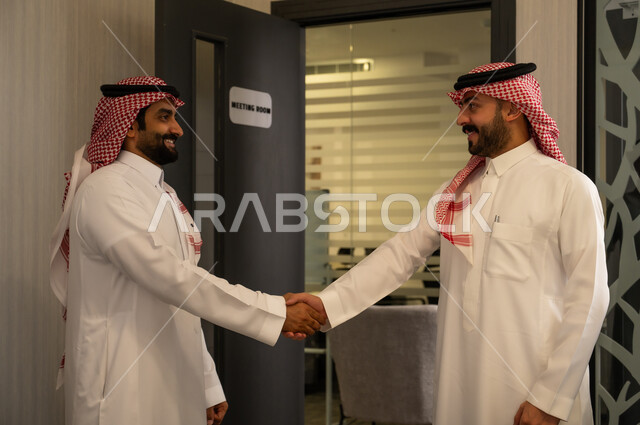 The concept of trust and building professional relationships, cooperation and partnership in the work environment, a successful business meeting, two Arab Gulf men from Saudi Arabia wearing white thobes and shemaghs shaking hands inside the office.