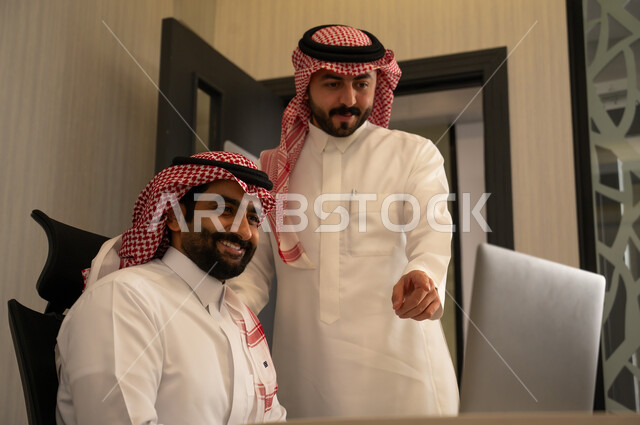 استخدام جهاز تقني حديث، الاشارة الى شاشة الحاسوب، جمع البيانات وحل المشكلات، اجتماع ومناقشة عمل في مقر الشركة، رجل عربي خليجي سعودي يرتدي الثوب و الشماغ  يقوم بالتحدث مع زميلة داخل المكتب، تطوير ودراسة المشاريع الجديدة في شركات السعودية