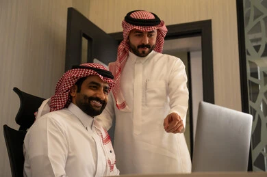 استخدام جهاز تقني حديث، الاشارة الى شاشة الحاسوب، جمع البيانات وحل المشكلات، اجتماع ومناقشة عمل في مقر الشركة، رجل عربي خليجي سعودي يرتدي الثوب و الشماغ  يقوم بالتحدث مع زميلة داخل المكتب، تطوير ودراسة المشاريع الجديدة في شركات السعودية