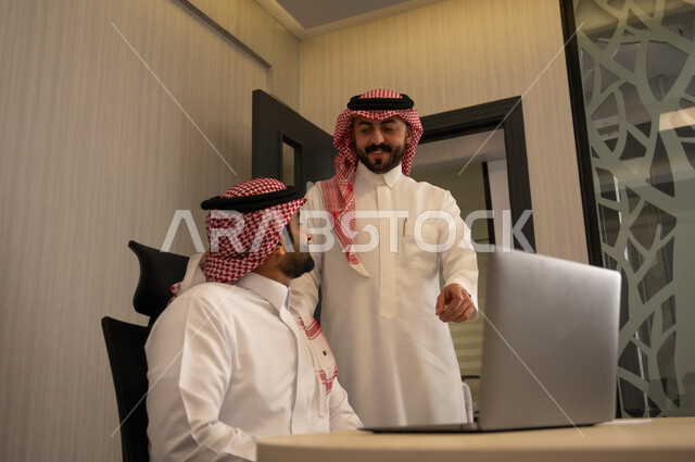 الاشارة الى شاشة الحاسوب، استخدام جهاز تقني حديث، جمع البيانات وحل المشكلات، اجتماع ومناقشة عمل في مقر الشركة، رجل عربي خليجي سعودي يرتدي الثوب و الشماغ  يقوم بالتحدث مع زميلة داخل المكتب، تطوير ودراسة المشاريع الجديدة في شركات السعودية
