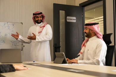 استخدام جهاز التابلت لتسجيل الملاحظات، ورشة عمل تدريبية مهنية، استخدام السبورة لسهولة الشرح، تطوير استراتيجيات العمل، تعلم اساليب العمل والتطوير المهني، رجل عربي خليجي سعودي يرتدي الثوب و الشماغ يقوم بتوضيح الخطوات لتحقيق الأهداف داخل مقر الشركة امام زميلة