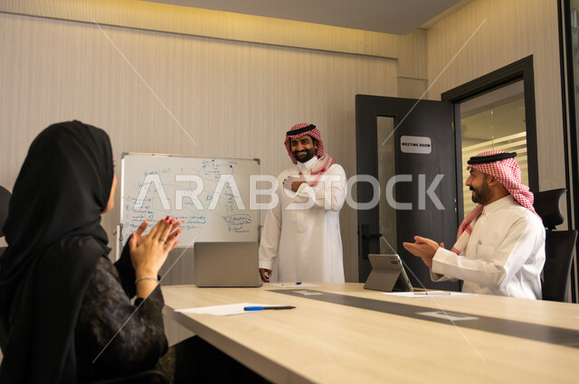 Cooperation and participation in completing tasks, developing plans and achieving company goals, cooperation and camaraderie among employees, an Arab Gulf Saudi team consisting of a man in a white thobe and shemagh and a veiled woman wearing an abaya, clapping and congratulating their colleague.