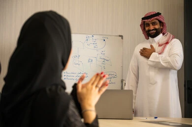 العمل الجماعي وروح الفريق، ايماءات التشجيع والتصفيق، ضمان فهم واضح وشامل للاعمال، استعمال السبورة لتوضيح العمليات والاجراءات المتبعة في الشركة، الاحترافية والتفاني في الإنجاز،امراة عربيه خليجيه سعودية محجبة تقوم بالتصفيق لزميلها