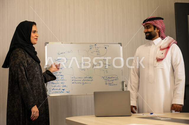الشرح على السبورة لضمان فهم واضح وشامل للاعمال، عقد اجتماع داخل مقر الشركة، رجل عربي خليجي اماراتي يرتدي الغترة والكندورة يقوم بتعليم اساليب العمل والتطوير المهني، التعاون والتفاهم بين الزملاء، التنظيم والتخطيط الاستراتيجي لتحقيق الأهداف،  امراه عربية محجبة ترتدي العباءة تقوم بتوضيح الخطوات لتحقيق الأهداف داخل مقر الشركة امام زميلها