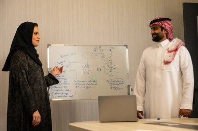 الشرح على السبورة لضمان فهم واضح وشامل للاعمال، عقد اجتماع داخل مقر الشركة، رجل عربي خليجي اماراتي يرتدي الغترة والكندورة يقوم بتعليم اساليب العمل والتطوير المهني، التعاون والتفاهم بين الزملاء، التنظيم والتخطيط الاستراتيجي لتحقيق الأهداف،  امراه عربية محجبة ترتدي العباءة تقوم بتوضيح الخطوات لتحقيق الأهداف داخل مقر الشركة امام زميلها