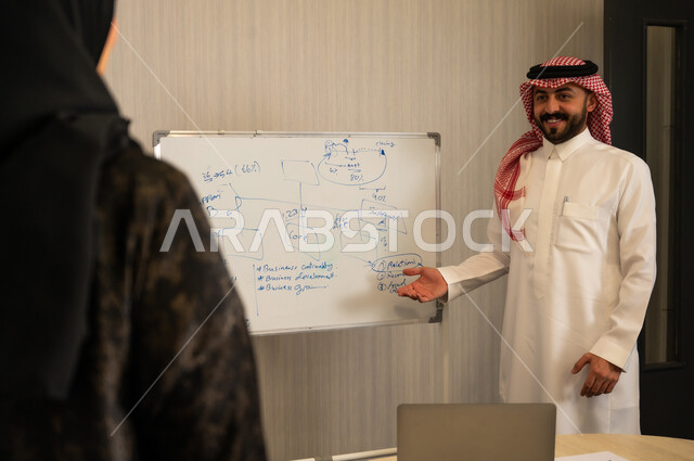 Developing work strategies, professional training workshops, learning work methods and professional development, using a whiteboard for easy explanation, an Arab Gulf Saudi man wearing a thobe and ghutrah is explaining the steps to achieve goals inside the company headquarters.