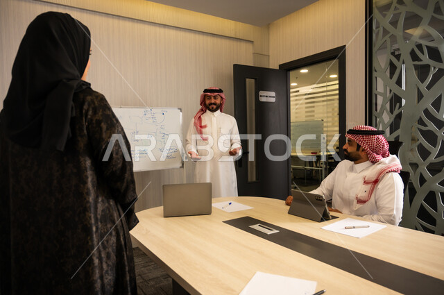 اجتماع فريق العمل لمناقشة مشروع، التخطيط الإداري داخل العمل, شرح أفكار ومخططات على السبورة, تفاعل مهني بين الموظفين داخل قاعة الاجتماعات، رجل عربي خليجي سعودي  يرتدي الثوب و الشماغ يقف ويشرح النقاط أمام زملائة