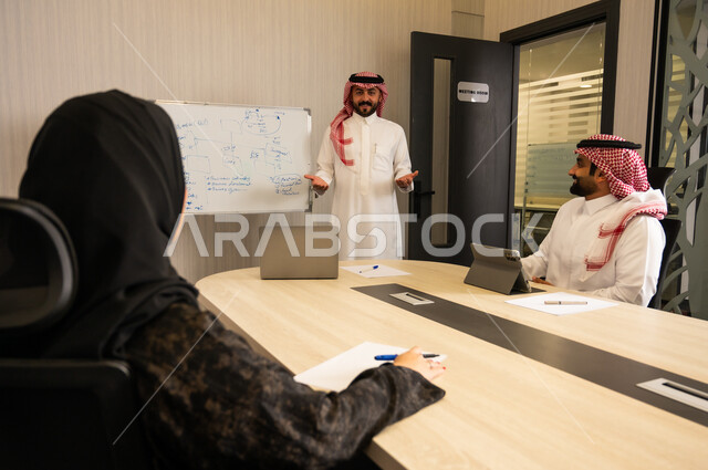 التخطيط الإداري داخل العمل, اجتماع فريق العمل لمناقشة مشروع, شرح أفكار ومخططات على السبورة, تفاعل مهني بين الموظفين داخل قاعة الاجتماعات، رجل عربي خليجي سعودي  يرتدي الثوب و الشماغ يقف ويشرح النقاط أمام زملائة