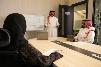 التخطيط الإداري داخل العمل, اجتماع فريق العمل لمناقشة مشروع, شرح أفكار ومخططات على السبورة, تفاعل مهني بين الموظفين داخل قاعة الاجتماعات، رجل عربي خليجي سعودي  يرتدي الثوب و الشماغ يقف ويشرح النقاط أمام زملائة