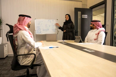 اجتماع عمل داخل قاعة الاجتماع, مناقشة الأفكار وتبادل الآراء المهنية, عرض مخطط وخطة عمل على السبورة البيضاء, موظفة عربية خليجية سعودية ترتدي العباءة والحجاب تشرح النقاط أمام الزملاء, رجلان عربيان خليجيان سعوديان يرتديان الزي التقليدي يتابعان العرض