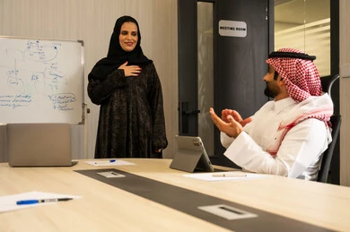 ايماءات السعادة و السرور، مفهوم التحفيز والدعم في بيئة العمل، إظهار التقدير أثناء عرض الأفكار, موظفة عربية خليجية سعودية تقوم بالشرح خلال اجتماع عمل, رجل عربي خليجي سعودي يرتدي الثوب التقليدي و الشماغ يقوم بالتصفيق