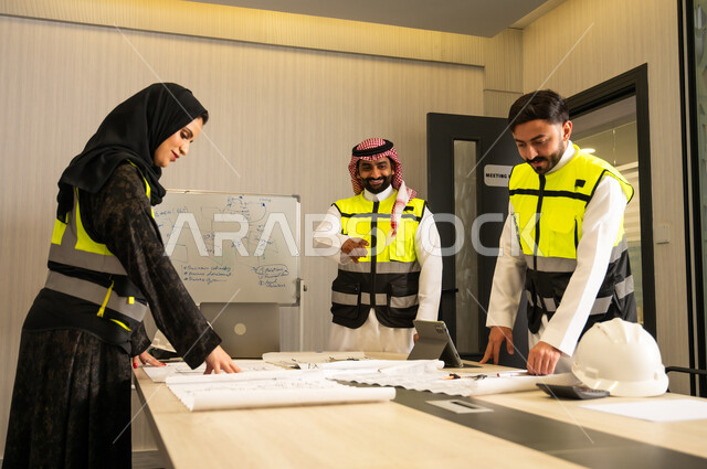 مراقبة وتنفيذ الخطط الهندسية المعتمدة، الشركات المختصة بورشات البناء والمقاولات، التخطيط ووضع المخططات المعمارية، مجموعة مهندسون عربيون خليجيون سعوديون يرتدون الثوب التقليدي و سترة الحماية يعملون على مناقشة مخطط الإنشاء وتحديد القياسات بدقة داخل مقر الشركة