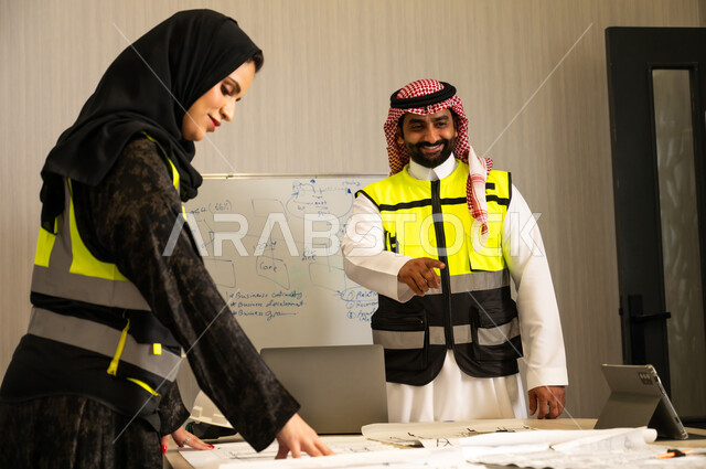 العمل الجماعي والتواصل المهني، مناقشة الأفكار وتبادل الآراء، التواصل الفعال وتنسيق المهام اليومية, موظف عربي خليجي سعودي يرتدي الثوب الابيض و الشماغ و سترة الحماية يقوم بشرح المهام لزميلته، الاشارة الى شيء ما