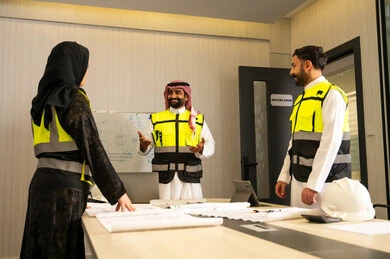 Supervising the progress of engineering work according to the established plans, companies specialized in construction workshops and contracting, planning and creating architectural designs, a group of Arab Gulf Saudi engineers wearing traditional attire and safety vests are discussing the construction plan and accurately determining the measurements inside the company's headquarters.