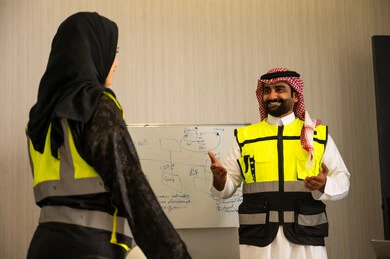 مناقشة الأفكار وتبادل الآراء، التواصل الفعال وتنسيق المهام اليومية, موظف عربي خليجي سعودي يرتدي الثوب الابيض و الشماغ و سترة الحماية يقوم بشرح المهام لزميلته ، العمل الجماعي والتواصل المهني