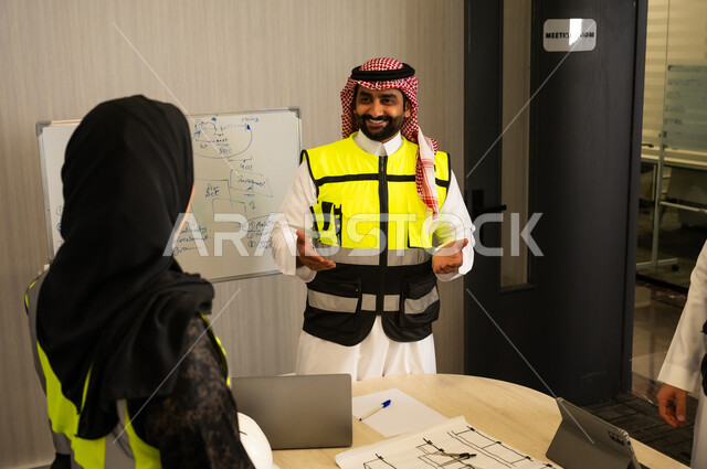التواصل الفعال وتنسيق المهام اليومية, موظف عربي خليجي سعودي يرتدي الثوب الابيض و الشماغ و سترة الحماية يقوم بشرح المهام لزميلته ، مناقشة الأفكار وتبادل الآراء، العمل الجماعي والتواصل المهني