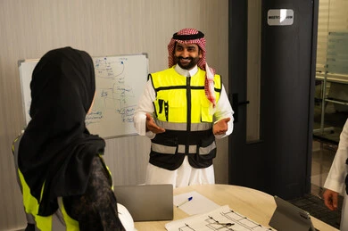 Effective communication and coordination of daily tasks, a Saudi Gulf Arab employee wearing a white thobe, a ghutrah, and a safety vest is explaining tasks to his colleague, discussing ideas and exchanging opinions, teamwork, and professional communication.