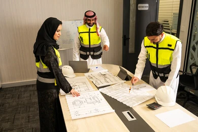 Monitoring the implementation of approved engineering plans, companies specialized in construction workshops and contracting, planning and developing architectural plans, a group of Arab Gulf Saudi engineers wearing traditional attire and safety vests are discussing the construction plan and accurately determining the measurements inside the company's headquarters.