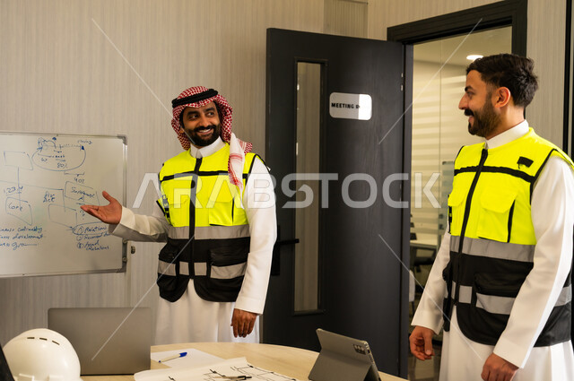 Using the whiteboard to clarify the processes and procedures followed, monitoring the implementation of the established engineering plans, companies specialized in construction workshops and contracting, planning and creating architectural designs, an Arab Gulf Saudi man wearing a thobe, ghutrah, and safety vest is explaining the work to his colleague.