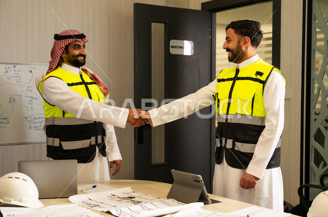 التعاون والاتفاق المهني داخل بيئة العمل, إبرام اتفاق بعد اجتماع عمل, مصافحة بالأيدي بتعابير الثقة والتفاهم, مهندسان عربيان خليجيان سعوديان يرتديان سترة السلامة يتصافحان داخل المكتب
