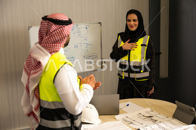 Gestures of encouragement and applause, ensuring a clear and comprehensive understanding of the work, using the board to clarify the processes and procedures followed in the company, teamwork and team spirit, professionalism and dedication to achievement, an Arab Gulf Saudi engineer wearing a thobe, ghutrah, and safety vest is applauding for his colleague.