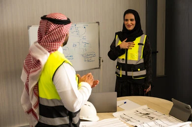 Gestures of encouragement and applause, ensuring a clear and comprehensive understanding of the work, using the board to clarify the processes and procedures followed in the company, teamwork and team spirit, professionalism and dedication to achievement, an Arab Gulf Saudi engineer wearing a thobe, ghutrah, and safety vest is applauding for his colleague.