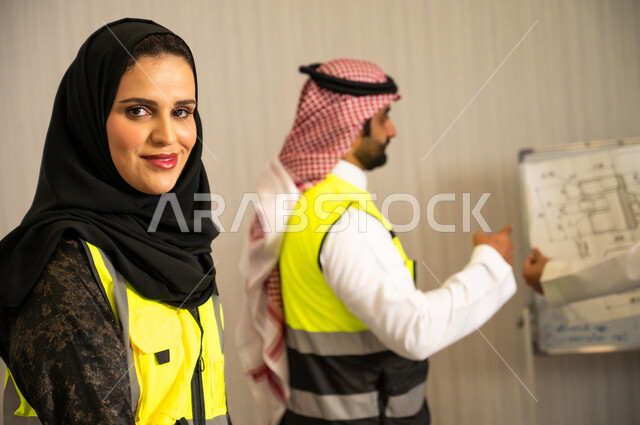 إدارة المشاريع الهندسية, مفهوم التواصل المهني, صورة مقربة لمهندسة عربية خليجية سعودية محجبة ترتدي سترة الحماية  تنظر الى الكاميرا بتعابير السرور، مهندس عربي خليجي سعودي يرتدي الشماغ و سترة السلامة يشرح المخطط الهندسي على لوحة عرض