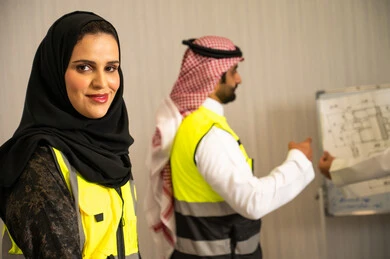 إدارة المشاريع الهندسية, مفهوم التواصل المهني, صورة مقربة لمهندسة عربية خليجية سعودية محجبة ترتدي سترة الحماية  تنظر الى الكاميرا بتعابير السرور، مهندس عربي خليجي سعودي يرتدي الشماغ و سترة السلامة يشرح المخطط الهندسي على لوحة عرض