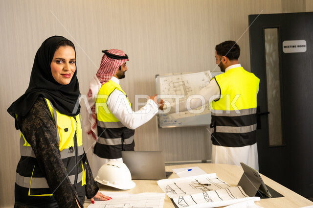 مفهوم التواصل المهني. إدارة المشاريع الهندسية, مهندسة عربية خليجية سعودية محجبة ترتدي سترة الحماية تنظر الى الكاميرا بتعابير السرور، مهندسان عربيان خليجيان سعوديان يرتديان الزي التقليدي و سترة السلامة يحملان المخطط الهندسي