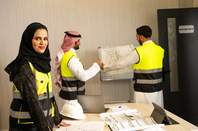 The concept of professional communication. Engineering project management, a Saudi Gulf Arab engineer wearing a hijab and a safety vest looking at the camera with a joyful expression, two Saudi Gulf Arab engineers wearing traditional attire and safety vests holding the engineering plan.