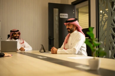 Unity and teamwork, integrating business with technology, accomplishing the required tasks, two Arab Gulf men from Saudi Arabia wearing traditional attire sitting in the office working on a laptop and a tablet, achieving the company's goals.