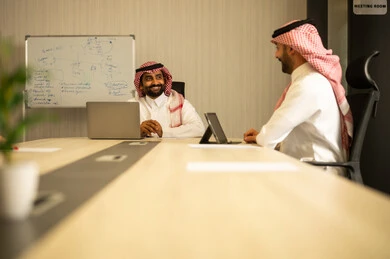 Integrating business with technology, accomplishing the required tasks, two Arab Gulf Saudi men wearing traditional attire sitting in the office working on a laptop and a tablet, achieving the company's goals, unity and teamwork.