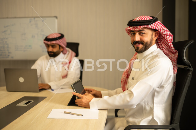 النظر للكاميرا بتعابير السرور،  إنجاز الأعمال المطلوبة، رجلان عربيان خليجيان سعوديان يرتديان الزي التقليدي يجلسانفي المكتب يعملان على الحاسب المحمول وجهاز التابلت،  دمج الأعمال مع التقنية و التكنولوجيا, تحقيق أهداف الشركة, الوحدة والعمل الجماعي