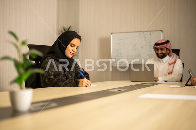 إدارة شؤون الأعمال وتنظيمها، استخدام جهاز تقني حديث ومتطور، مهنة ووظيفة ادارية مكتبية، موظف عربي خليجي سعودي يرتدي الثوب التقليدي يقوم بالعمل على الحاسوب المحمول داخل المكتب، الشركات والمكاتب في المملكة العربية السعودية، امراه عربية خليجيه سعودية محجبة ترتدي العباءة تقوم بكتابة و تدوين الملاحظات