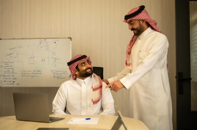 العمل على الخطة لتحقيق اهداف الشركة، دمج العمل مع التقنية والتكنولوجيا، تطور الشركات السعودية، متابعة مشاريع الأعمال، رجل عربي خليجي سعودي يرتدي الثوب و القماغ يجلس امام الحاسب المحمول يقوم بمناقشة زميلة، الاشارة الي شاشة الابتوب