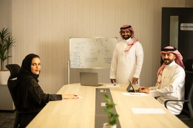 Looking at the camera with expressions of joy, using a tablet to take notes, ensuring a clear and comprehensive understanding of the work, using a whiteboard to clarify the processes and procedures followed in the company, teamwork and collaboration, an Arab Gulf Saudi man wearing a white thobe and a ghutrah explaining the strategy to his colleagues.