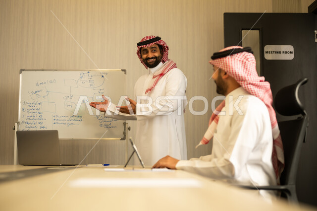 ضمان فهم واضح و شامل للاعمال، استعمال السبورة لتوضيح العمليات والاجراءات المتبعة في الشركة، العمل الجماعي و التعاون، رجل عربي خليجي سعودي يرتدي الثوب الابيض و الشماغ يقوم بشرح الاستيراتيجية لزميلة