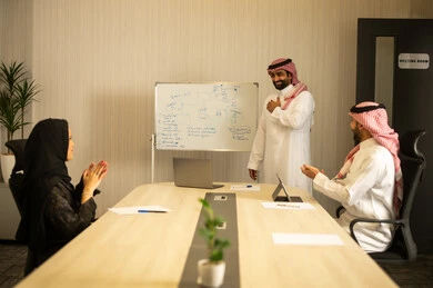 Developing plans and achieving the company's goals, cooperation and love among employees, collaboration and participation in completing tasks, a Gulf Arab Saudi team consisting of a man in a white thobe and ghutrah and a woman in a hijab wearing an abaya, they are applauding and congratulating their colleague.